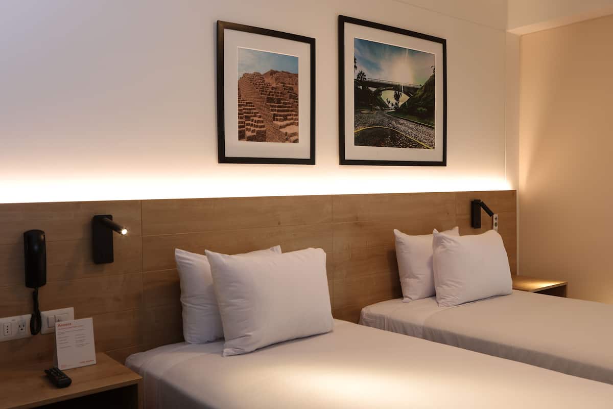 A hotel room with two beds, a wooden headboard, a nightstand with a phone, and framed pictures on the wall.