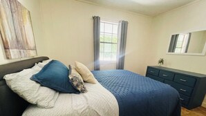 Ein Schlafzimmer mit Bett, Kommode, Fenster mit Vorhängen und Spiegel an der Wand.