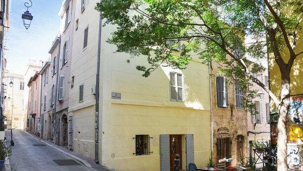 Une ruelle étroite pavée, bordée d’immeubles, avec un arbre qui offre de l’ombre et des tables et des chaises disposées à l’extérieur pour créer un espace de détente.