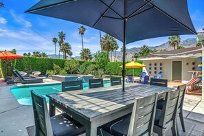 A poolside area with a wooden table and chairs, a large umbrella, and a view of mountains in the background.