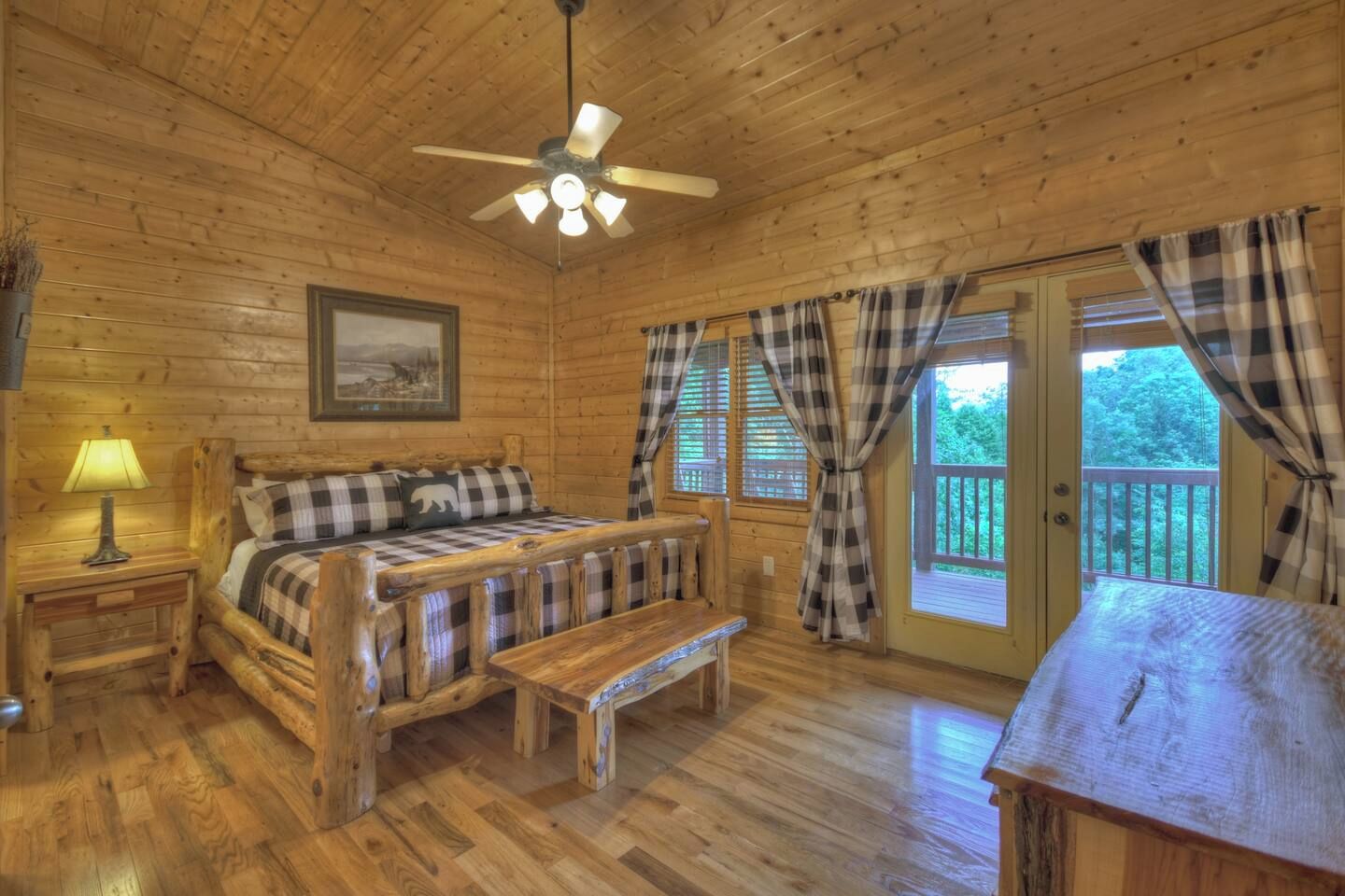 Une pièce de style cabane en bois, comprenant un lit, un banc, un ventilateur de plafond et une fenêtre avec des rideaux.