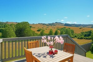 Um deck de madeira com mesa e cadeiras, um vaso de flores cor-de-rosa, e uma vista panorâmica de colinas e árvores.