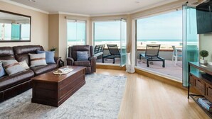 A spacious living room with a large window overlooking a balcony, a leather sofa, a coffee table, and a television. - Ocean Front Beach Get Away w/ AC! (San Diego)
