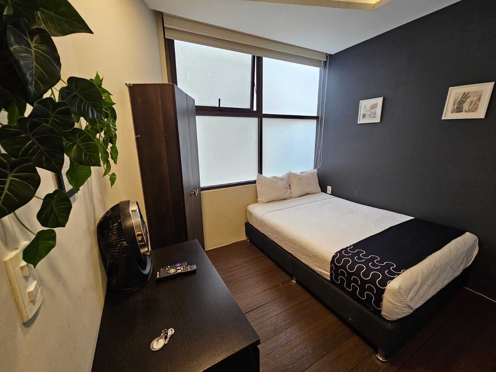 A bedroom with a bed, a window, a plant, a desk with a remote control, and a wall-mounted light.