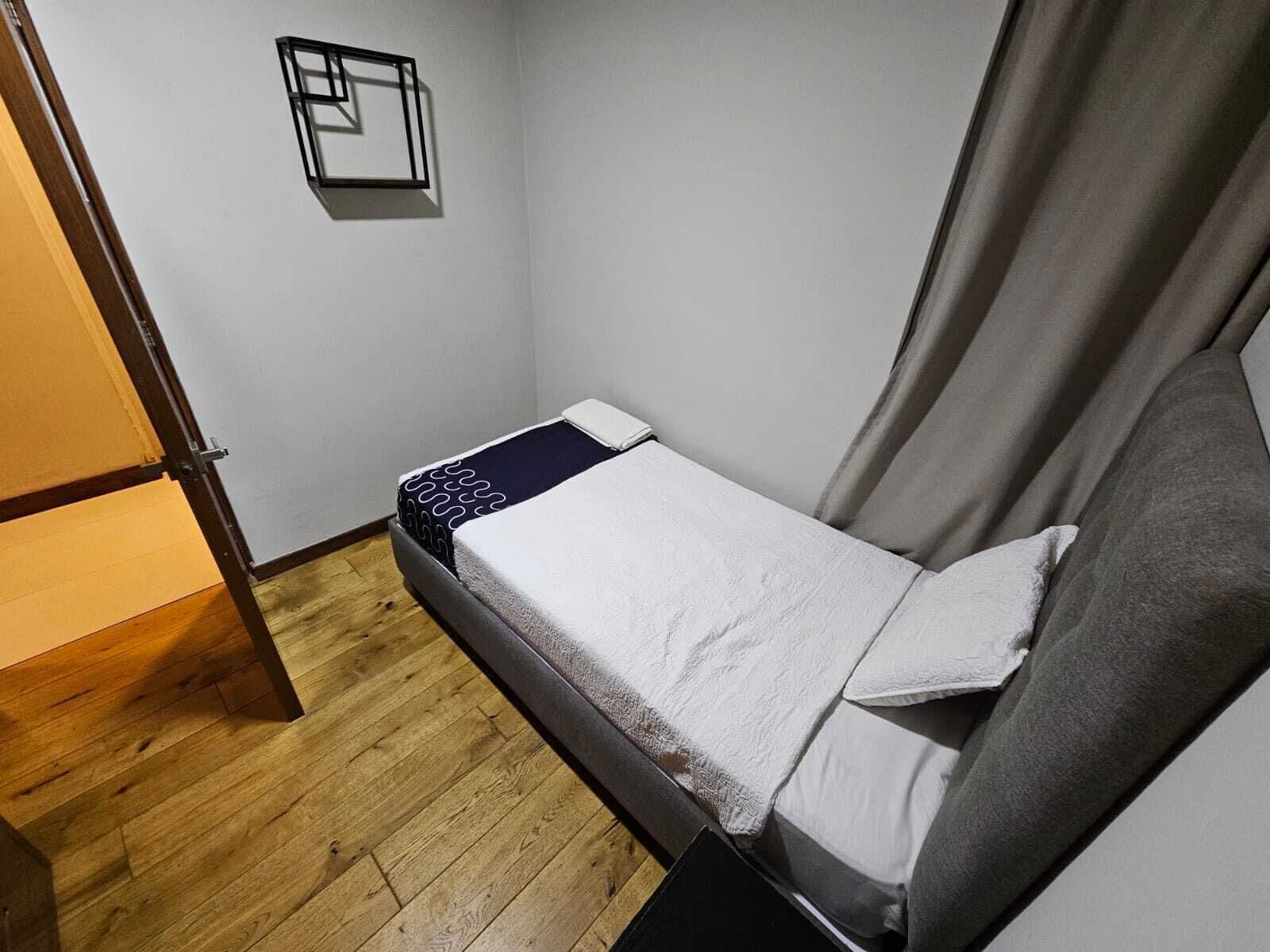 A small, single-bed room with a wooden floor, a grey headboard, and a white bedspread.