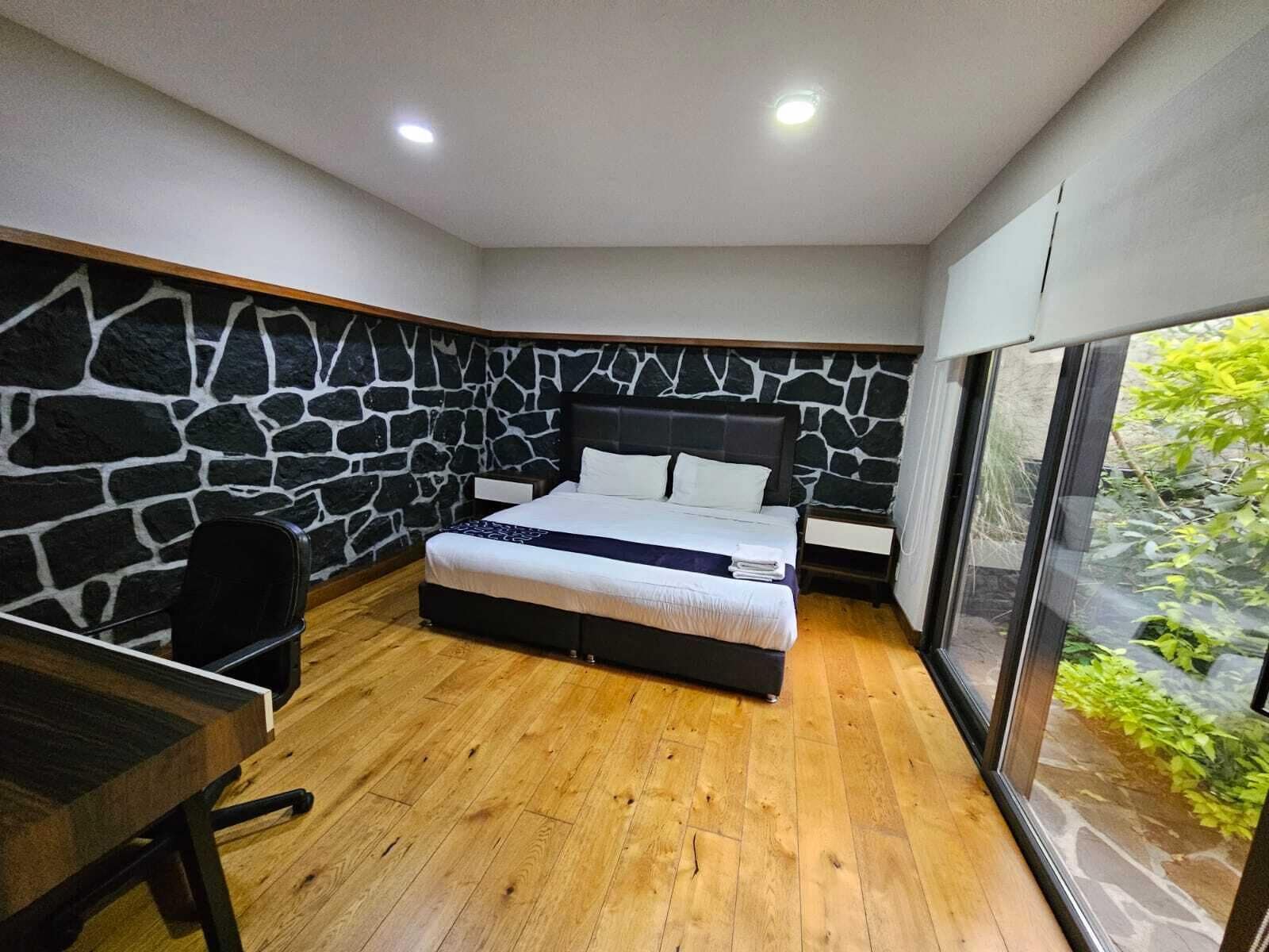 A bedroom with a stone wall, a bed, a desk, and a large window.