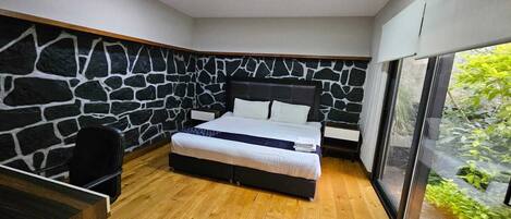 A bedroom with a stone wall, a bed, a desk, and a large window.