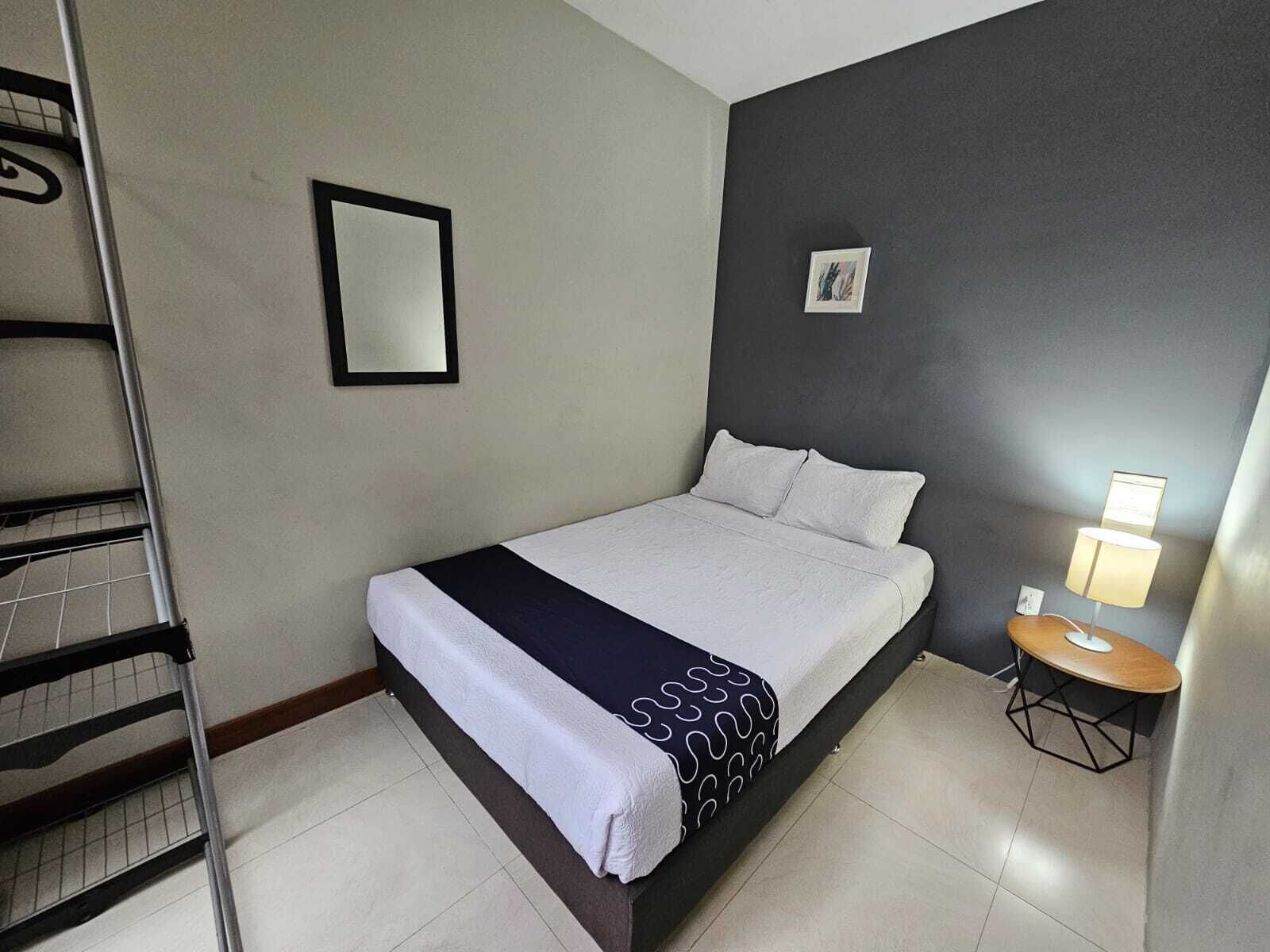 A single bed with a white and black bedspread, a small bedside table with a lamp, a framed picture on the wall, and a metal ladder against the wall.