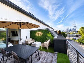 Un patio al aire libre con una mesa y sillas, un paraguas grande, un letrero de neón y un horizonte urbano al fondo.