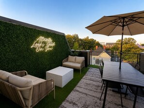 Un patio al aire libre con un muro cubierto de verde, un letrero de neón, un sofá, una mesa y una sombrilla.