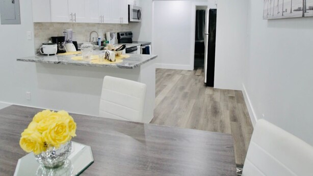 A modern kitchen with white cabinets, a microwave, and a refrigerator. There is a dining area with a wooden table and a vase of yellow flowers.