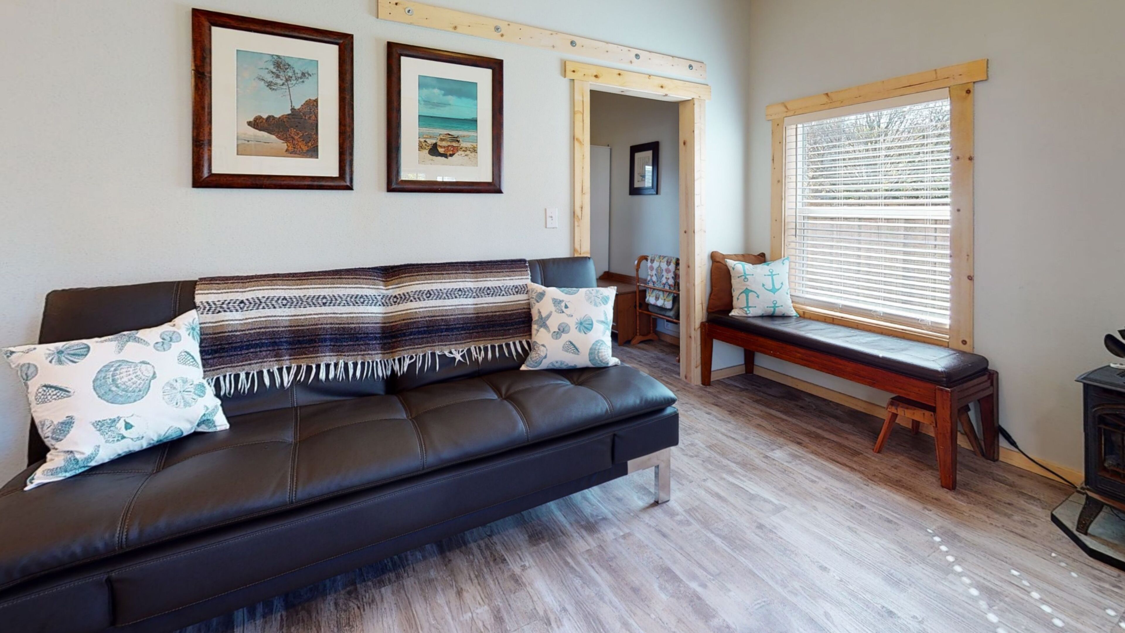 A living room with a dark leather sofa, a bench, and a window with blinds. There are two framed pictures on the wall.