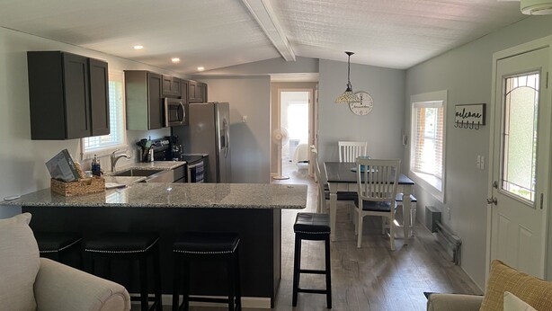 A modern kitchen with dark cabinets, a stainless steel refrigerator, and a granite countertop. There is a dining area with a white table and chairs, and a living room with a beige sofa and a coffee table.