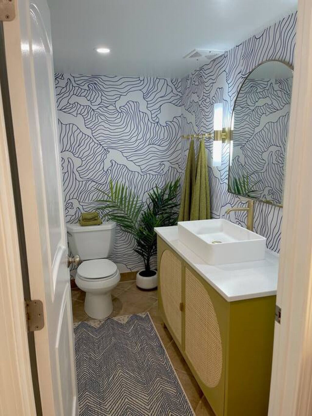 Une salle de bain avec des toilettes blanches, un lavabo blanc et un meuble vert. Il y a un miroir au-dessus du lavabo et une plante en pot à côté des toilettes.
