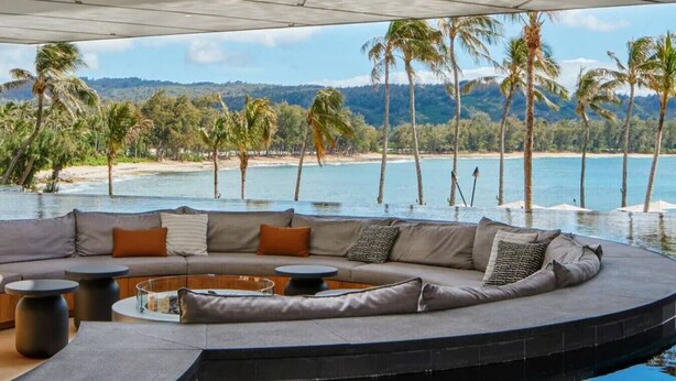 A modern outdoor seating area with a curved sofa, round tables, and a fire pit, overlooking a beach and palm trees. - SEArider ONE at Turtle Bay (2 bedroom / 2 bath) (Kahuku)