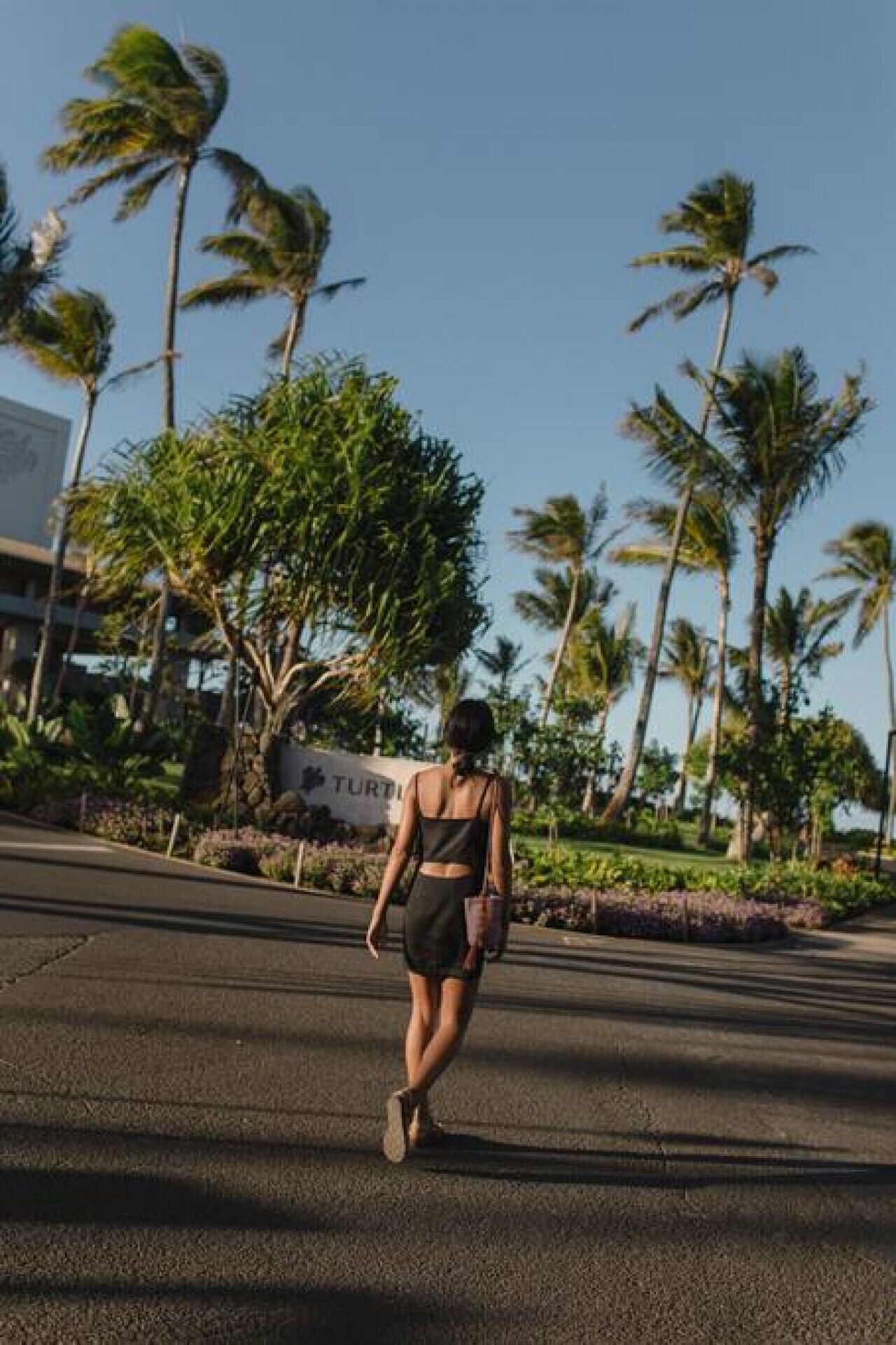 A person walking on a paved path with palm trees and a sign that reads "TURING" in the background.