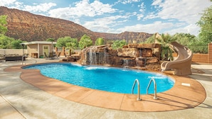Una piscina con una cascada, rodeada por un patio con mobiliario exterior y un telón de fondo montañoso.
