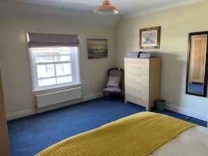 Ein Schlafzimmer mit Bett, Heizkörper, einem Fenster mit Vorhängen, einem Sessel, einem Schminktisch und einem Bild an der Wand.