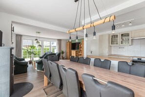 A modern kitchen with wooden cabinets, a dining area with a long table and chairs, and a view of greenery outside. - House Sommerwind iii near the North Sea with Fireplace, Wi-Fi, Garden, Terrace & BBQ (Tating)