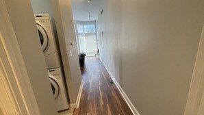Un couloir doté d’un plancher en bois franc, un espace buanderie avec des lave-linge et des sèche-linge superposés, et une porte donnant sur une pièce avec une fenêtre.