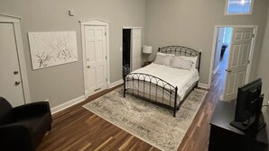 Une chambre avec un lit, un bureau et un téléviseur. Il y a deux portes et une fenêtre.