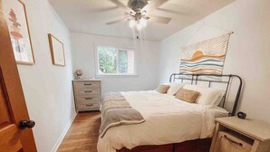 Une chambre à coucher comprenant un lit, une table de chevet, une commode, un meuble à tiroirs, un ventilateur de plafond et une fenêtre.