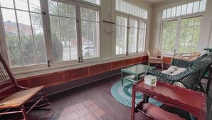 Une pièce dotée de grandes fenêtres, d’un plancher en bois, d’un fauteuil à bascule, d’une table et de chaises en osier.