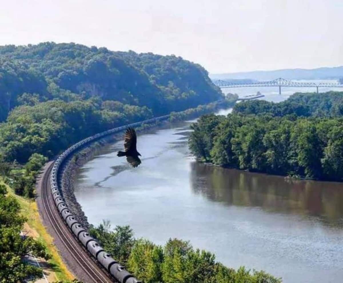 Ein Zug fährt auf einer gewundenen Strecke entlang, parallel zu einem Fluss, mit einer Brücke im Hintergrund.