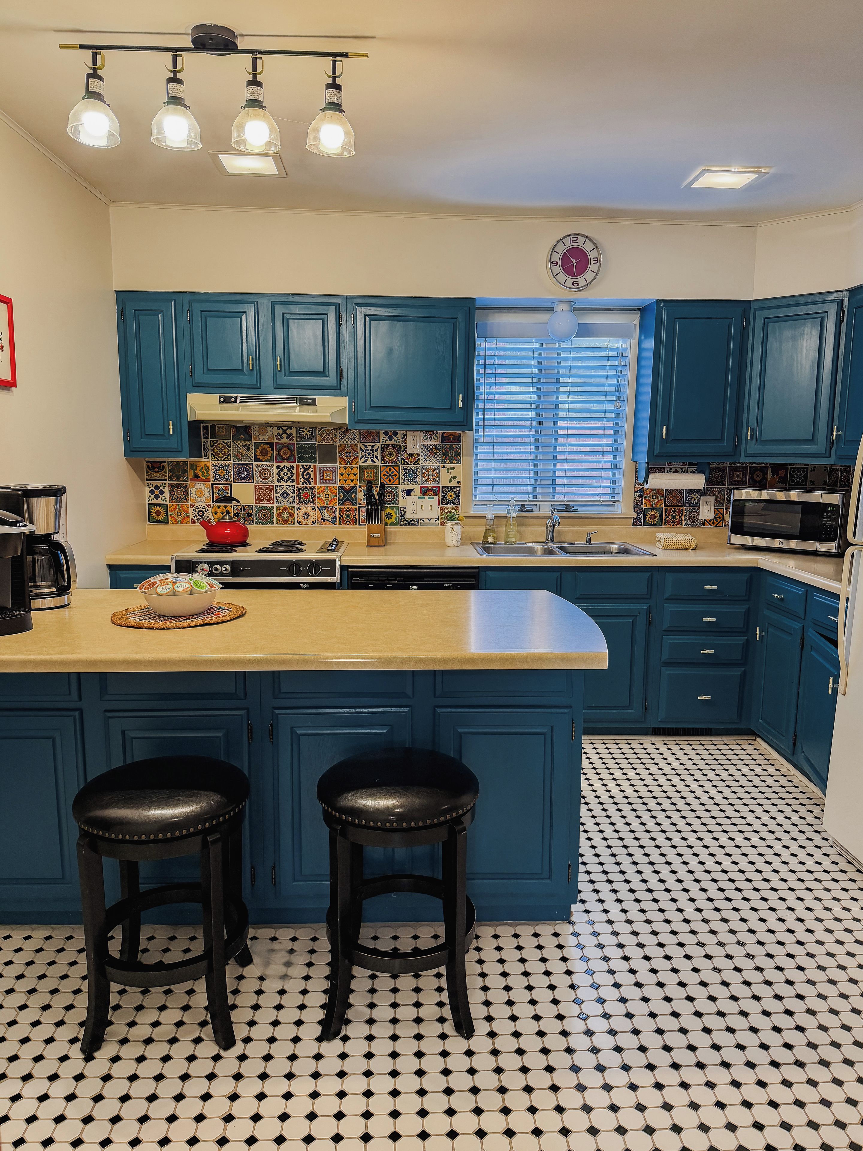 A kitchen with blue cabinets, a black and white tiled floor, a central island with stools, and a window with blinds.
