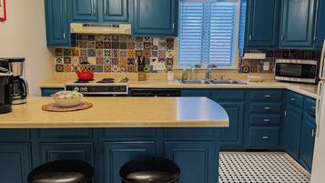 A kitchen with blue cabinets, a black and white tiled floor, a central island with stools, and a window with blinds.