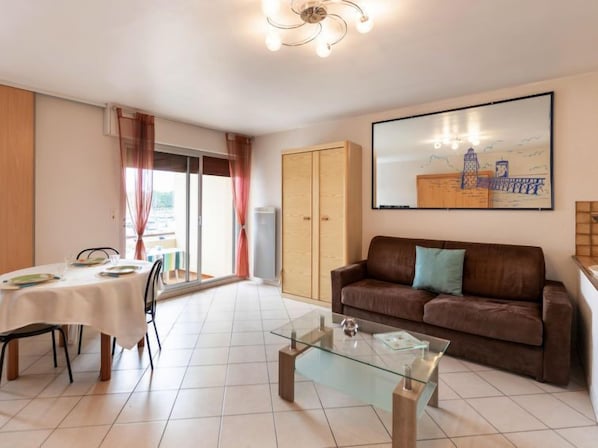 A living room with a glass coffee table, a brown sofa with a pillow, a dining table with plates and glasses, and a sliding glass door leading to an outdoor area. - le Grand Foc by Interhome (Capbreton)