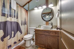 Ein Badezimmer mit einem Holzkonsolenunterschrank, einer weißen Toilette, einem Duschvorhang mit Tiermotiven und einem runden Spiegel mit Beleuchtung darüber.