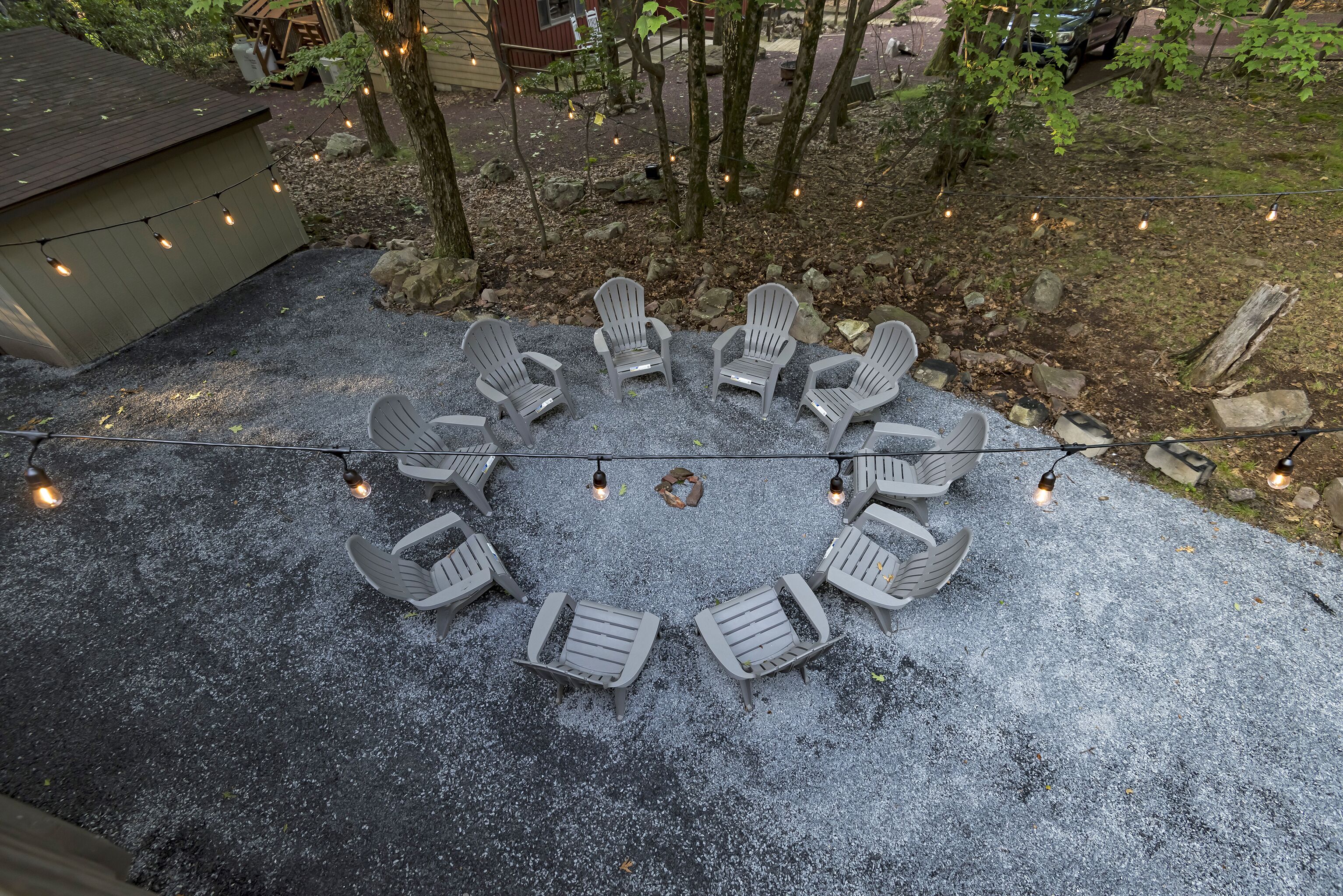 Disposizione circolare di poltrone Adirondack attorno a un braciere su un patio sterrato, circondato da alberi e luci decorative.