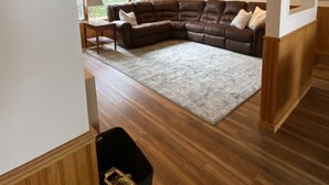 A spacious living room with a large L-shaped sectional sofa, a wooden floor, and a view through a window. - Peaceful Lakefront Home for Rent on Side Lake chain of lakes! (Side Lake)