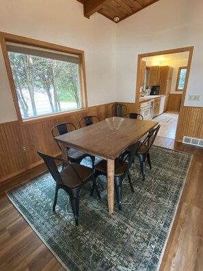 A dining area with a wooden table and chairs, a patterned rug, and large windows with a view of trees.