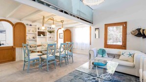 Amplia sala de estar con una mesa de centro de vidrio, un sofá con cojines decorativos, y un comedor con sillas azules y una mesa de madera.