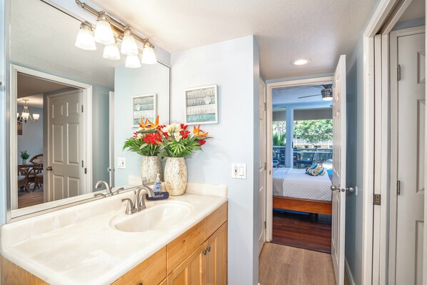 A bathroom with a large mirror, a sink, and a vanity. There are two framed signs on the wall and a vase with flowers on the vanity. - Maui Banyan G202 - King Bed, Ocean View, Across from Kamaole Beach (Kihei)