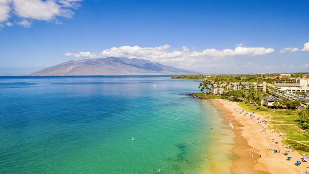 A coastal beach with clear turquoise waters, a sandy shore, and a mountain in the background. - Maui Banyan G202 - King Bed, Ocean View, Across from Kamaole Beach (Kihei)