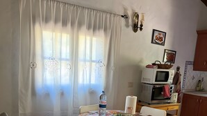 A dining area with a table covered in a patterned tablecloth, surrounded by white chairs, a kitchen area with stacked appliances, and a window with curtains. - Country Cabin "Finca Nim 2" on a Farm with Mountain View, Pool & Wi-Fi (Mogán)