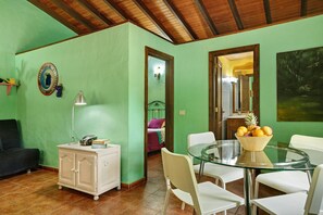 Une pièce avec un mur vert, une armoire blanche, une table en verre avec des chaises, et un lit à l’arrière-plan.