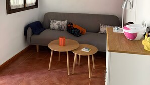A living room with a sofa, two small wooden tables, a coffee table, a pink and white speaker, a shelf with decorative items, and a window with curtains. - Country Cabin "Finca Nim 2" on a Farm with Mountain View, Pool & Wi-Fi (Mogán)