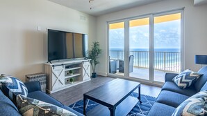 Ett vardagsrum med en platt-TV, en takfläkt, en stor skjutdörr i glas som leder ut till en balkong med utsikt över havet och en soffgrupp.