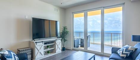 Uma sala de estar com televisão de tela plana, ventilador de teto, porta de vidro deslizante para uma varanda com vista para o oceano e um conjunto de sofás.