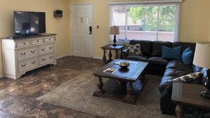 Una sala de estar con una televisión sobre un mueble bajo, un sofá con cojines, una mesa de centro y una ventana con persianas.