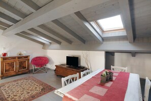 Een kamer met een schuine plafond, een houten tafel met een rode tafelkleed, een televisie op een houten dressoir, en een rode stoel.