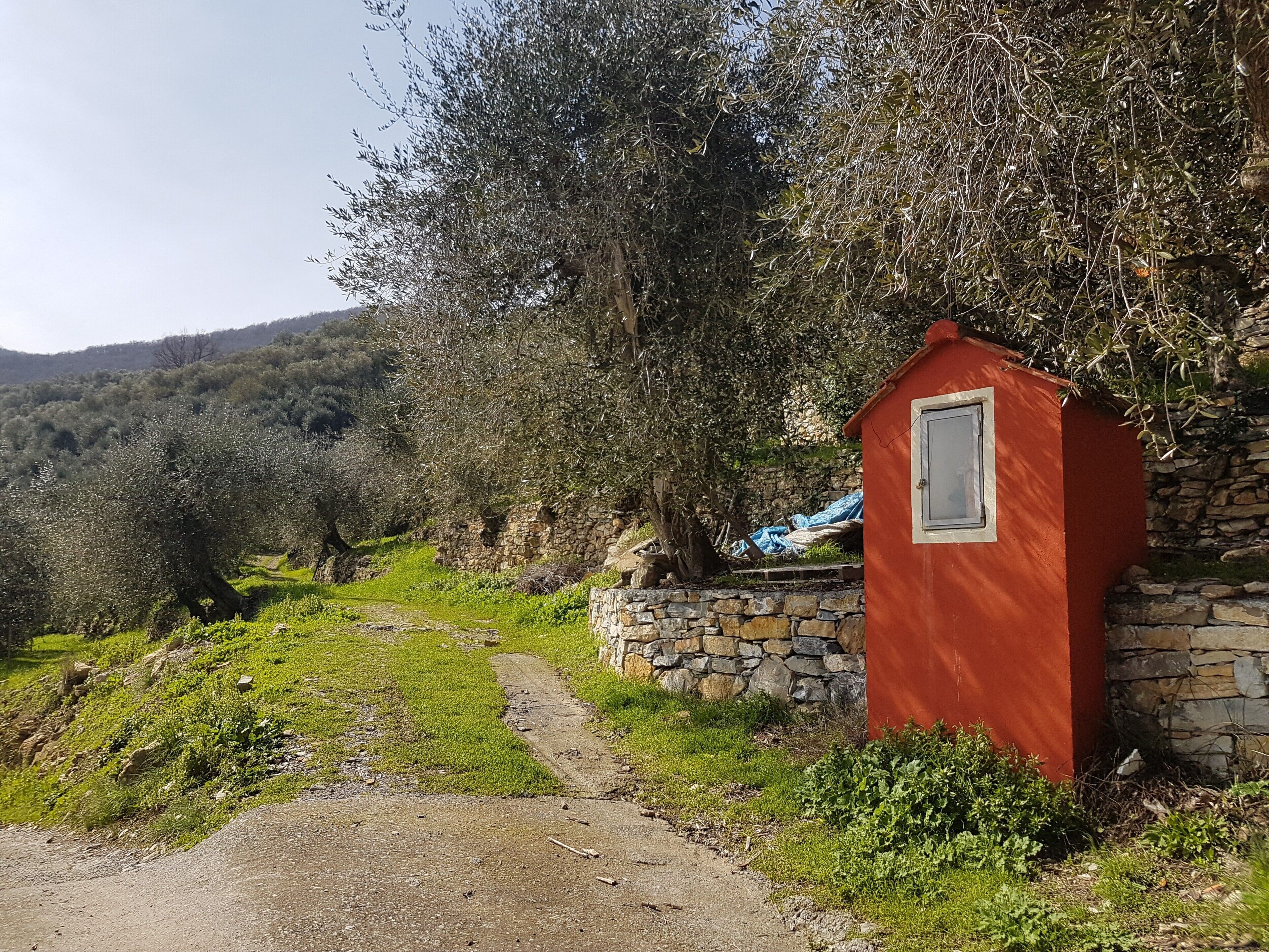 Een rood gebouw met een raam, omringd door stenen muren en olijfbomen.