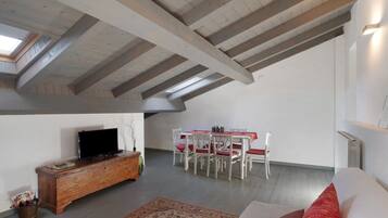 A loft-style room with exposed wooden beams, a television on a wooden cabinet, a dining table with chairs, and a radiator.