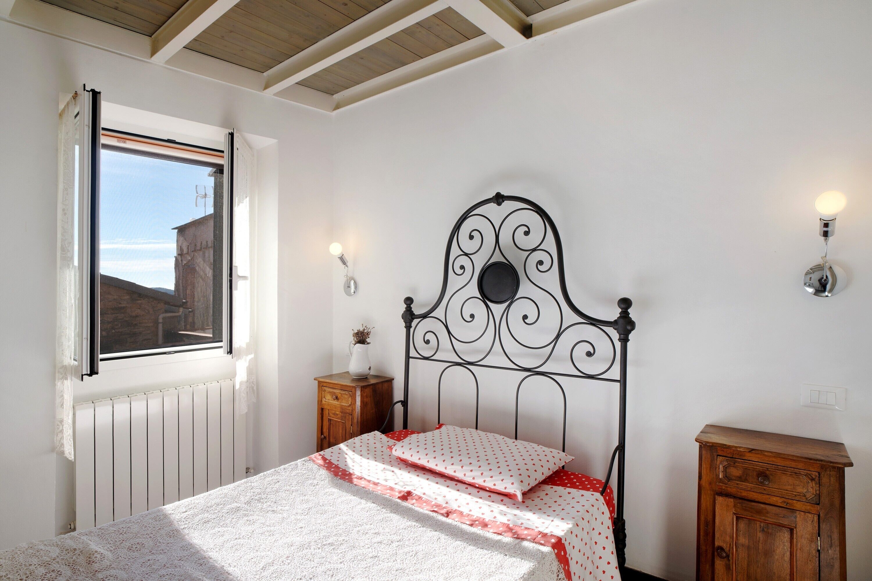 Una camera da letto con un letto, una finestra, un termosifone, un mobiletto in legno e luci a parete.