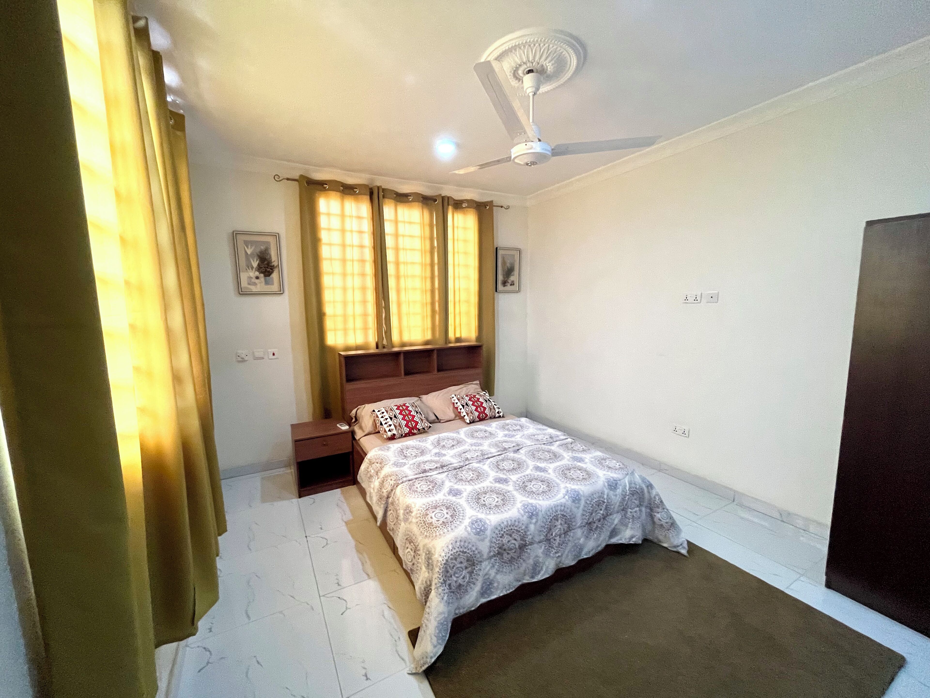 Quarto com cama, mesa de cabeceira, ventilador de teto, janela com cortinas e porta.