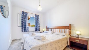 Un dormitorio con dos camas, cabeceros de madera, una mesita de noche con lámpara, un espejo y una ventana con cortinas estampadas.